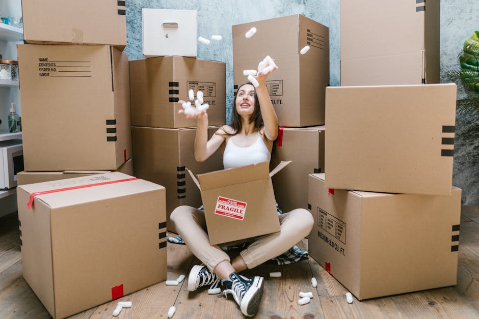 A person’s hand reaching inside a partially open cardboard moving box on a light wooden floor, surrounded by several similar packed boxes of varying sizes, some sealed with tape. The boxes are made 