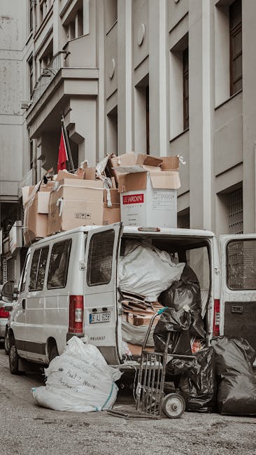 A white commercial van parked on a city street with its rear doors open, revealing a load of packing materials, cardboard boxes, and plastic bags filled with belongings for a house relocation. Several