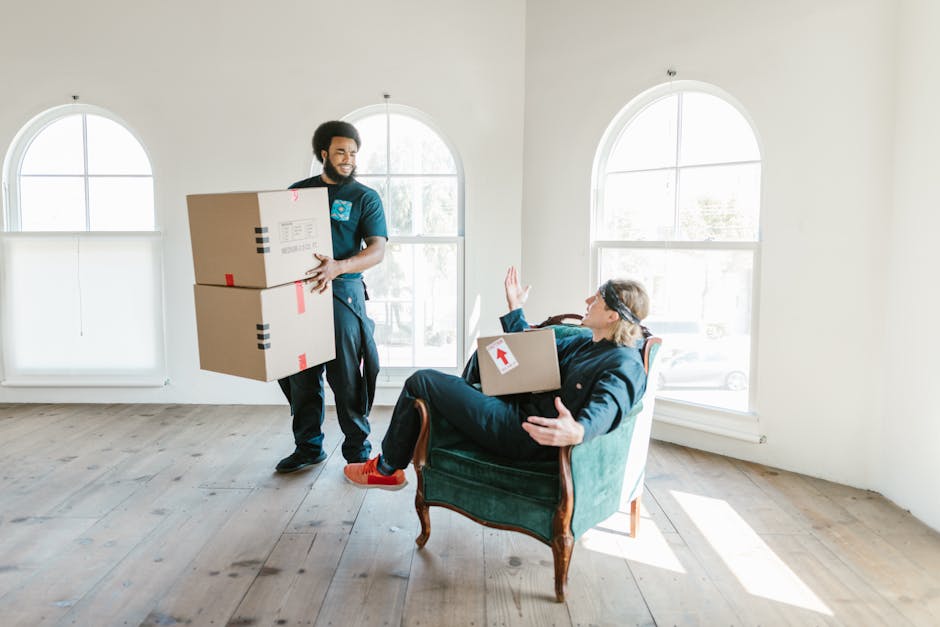 A person wearing light-colored clothing and white socks is seen in a room, carefully lifting and stacking a cardboard box onto a larger filled box amidst several other packed boxes. The boxes are plai